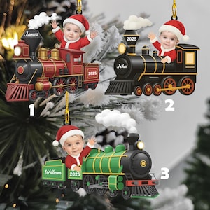 May include: Three Christmas ornaments shaped like toy trains, each featuring a baby's photo wearing a Santa hat and suit. The trains are red, black, and green, with the names "James", "John", and "William" on them, along with the year "2025".