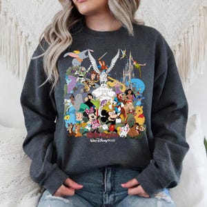 Puede incluir: Sudadera gris oscuro con un colorido collage de personajes de Disney, incluyendo Mickey y Minnie Mouse, Peter Pan y Dumbo. Las palabras "Magic Kingdom" y "Walt Disney World" est&aacute;n impresas en la parte inferior.