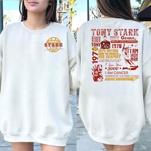 May include: Cream-colored sweatshirt with a front chest graphic of the Stark Industries logo and text. The back features a red graphic with the text "Tony Stark" and other phrases, including "I am Iron Man."