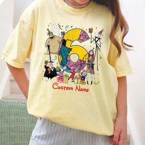May include: A pale yellow short-sleeve t-shirt featuring a large graphic of the number six surrounded by characters from The Nightmare Before Christmas, including Jack Skellington, Sally, and Oogie Boogie, with the red text Custom Name below.