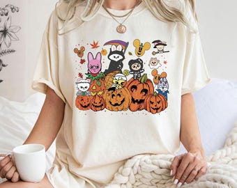 May include: Cream-colored t-shirt featuring a Halloween-themed graphic. The design includes several carved pumpkins with various cartoon characters. The characters include a bunny, a grim reaper, and other spooky figures. The shirt has short sleeves and a relaxed fit.