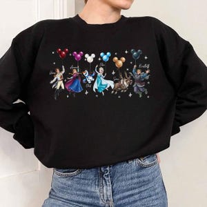 May include: Black sweatshirt featuring a graphic of characters from the movie Frozen, including Elsa, Anna, Olaf, and Kristoff. Each character is holding a Mickey Mouse-shaped balloon. The names of the characters are printed above them.