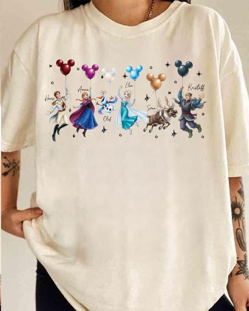 May include: Cream-colored t-shirt featuring a colorful graphic of characters from a popular animated movie. The characters are holding balloons in various colors. Names are written above the characters. The design includes stars.
