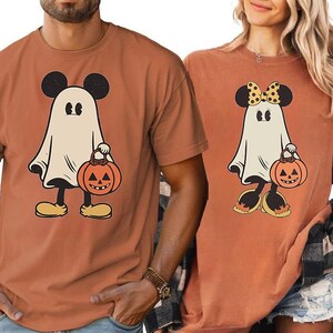 May include: Two rust-colored t-shirts featuring Halloween-themed graphics. The left shirt displays a ghost character with mouse ears holding a jack-o'-lantern. The right shirt shows a ghost character with a polka-dot bow and mouse ears holding a pumpkin.