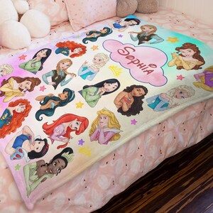 May include: A white and pastel-colored blanket featuring illustrations of various princesses. The blanket has a cloud-shaped text bubble with the name "Sophia" in pink. The princesses are in various poses, surrounded by stars and crowns.