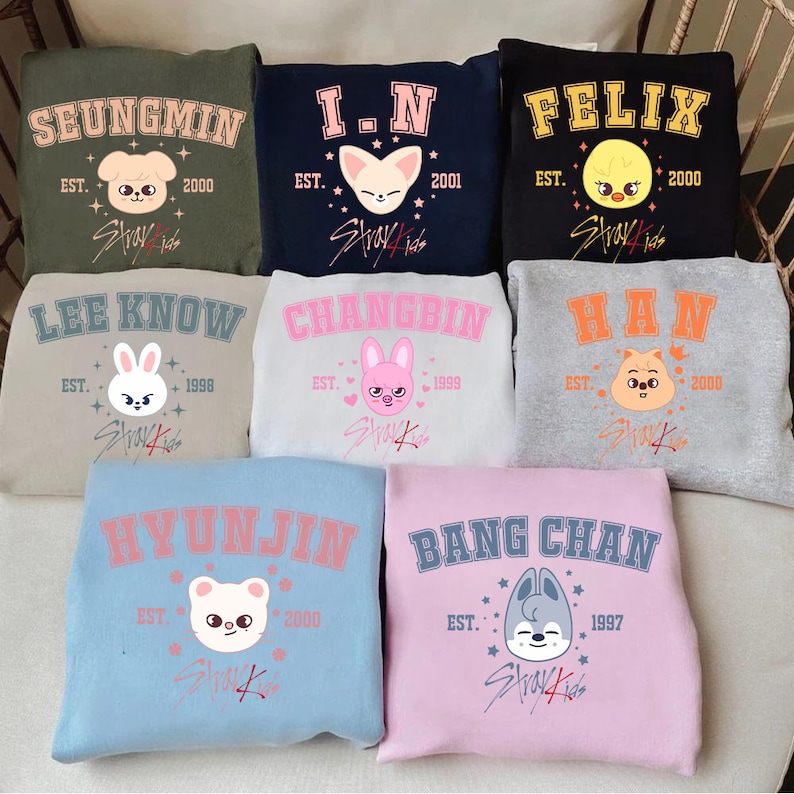 May include: Nine crewneck sweatshirts in different colours, each with a name in a collegiate font. Each sweatshirt has a cartoon animal graphic and the text "Stray Kids". The sweatshirts are for sale.