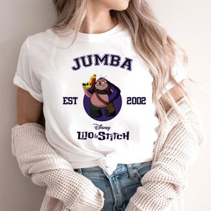 May include: White t-shirt featuring a graphic of Jumba from Disney's Lilo & Stitch. The design includes the text "JUMBA", "EST 2002", and the Disney Lilo & Stitch logo. Jumba is depicted holding a yellow and red weapon.