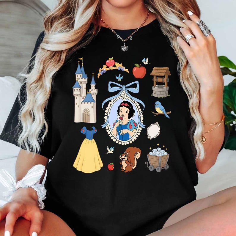 May include: Black t-shirt featuring a colorful graphic of Snow White and various related elements. The design includes a castle, dress, apple, well, and a squirrel. The shirt has a crew neck and short sleeves.