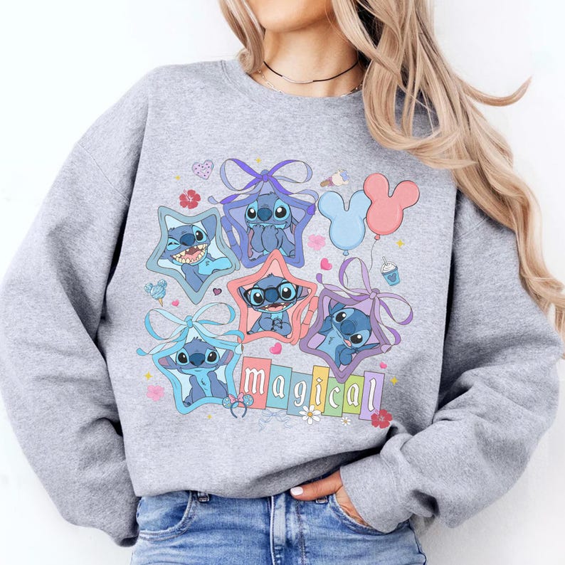 Może przedstawiać: Jasnoszara bluza z kolorową grafiką postaci Stitch z Lilo & Stitch. Projekt zawiera Stitch w gwiazdkowych ramkach, kokardkach i słowo "magical". Balony i kwiatowe akcenty dopełniają projekt.