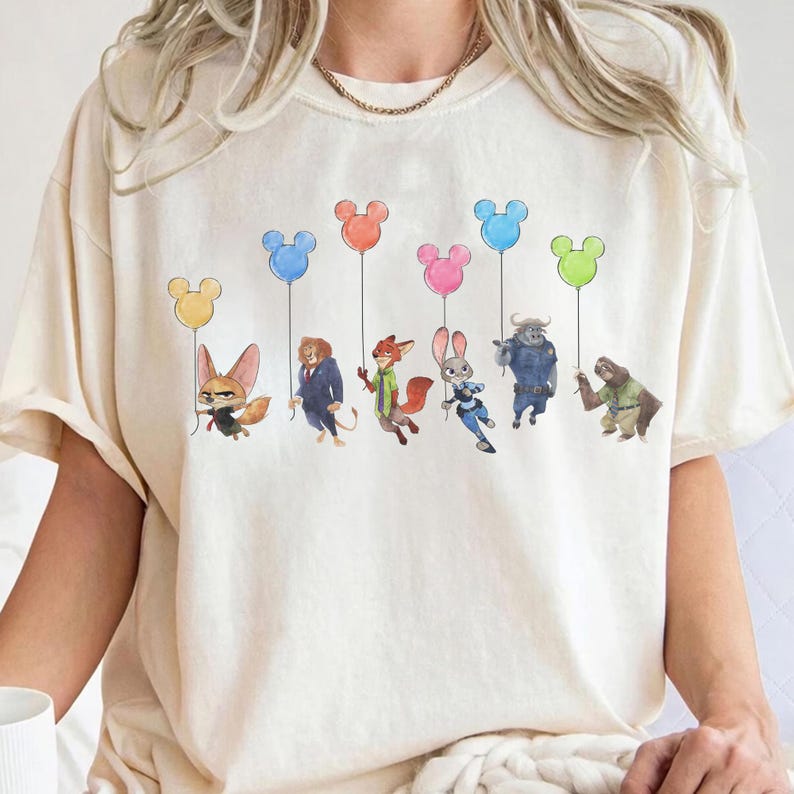 May include: Cream-colored t-shirt featuring a whimsical illustration of cartoon characters holding Mickey Mouse-shaped balloons in various colors. The characters include a fox, lion, rabbit, and others. The design is centered on the shirt.