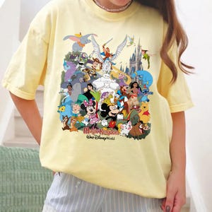 Puede incluir: Una camiseta amarilla clara con un gr&aacute;fico colorido de varios personajes de Disney, incluyendo Mickey Mouse, Minnie Mouse y Winnie the Pooh. El texto "Magic Kingdom Walt Disney World" est&aacute; impreso debajo.