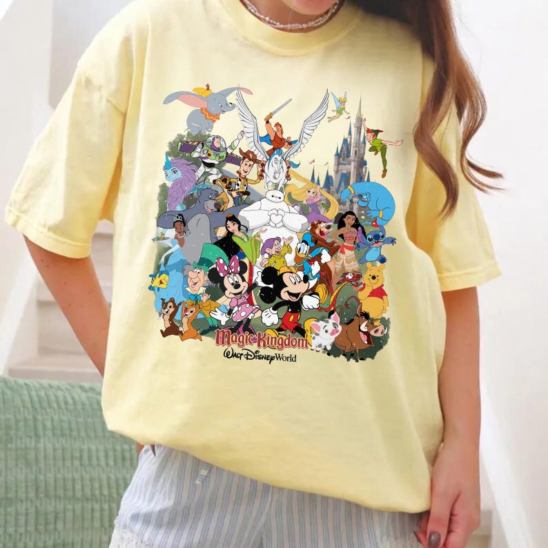 Puede incluir: Una camiseta amarilla clara con un gr&aacute;fico colorido de varios personajes de Disney, incluyendo Mickey Mouse, Minnie Mouse y Winnie the Pooh. El texto "Magic Kingdom Walt Disney World" est&aacute; impreso debajo.
