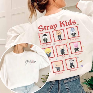 May include: White sweatshirt featuring the text "Stray Kids" in black script on the front and a design on the back. The back design includes the words "Stray Kids" in red, along with cartoon character illustrations in red postage stamp frames.