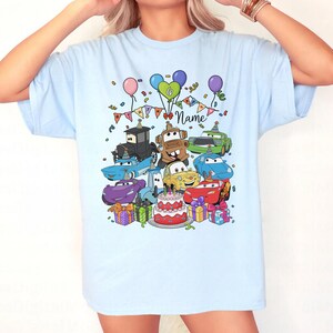 Peut inclure: T-shirt bleu clair avec un motif color&eacute; de voitures de dessins anim&eacute;s, un g&acirc;teau d'anniversaire, des ballons et des confettis. Le motif comprend le mot "Name" en &eacute;criture manuscrite, id&eacute;al pour la personnalisation. Un design festif et amusant.