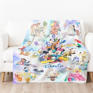 Op de afbeelding: Een witte fleece deken met een kleurrijk ontwerp van Disney-figuren, waaronder Mickey en Minnie Mouse, Donald Duck en personages uit Toy Story, Cars en Frozen. De naam "DAVID" is in blauw geschreven.