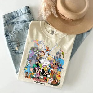 Puede incluir: Camiseta color crema con un gr&aacute;fico colorido de personajes de Disney, incluyendo Mickey y Minnie Mouse, Dumbo y Buzz Lightyear. La camiseta se combina con vaqueros azul claro, un sombrero beige y un collar plateado.