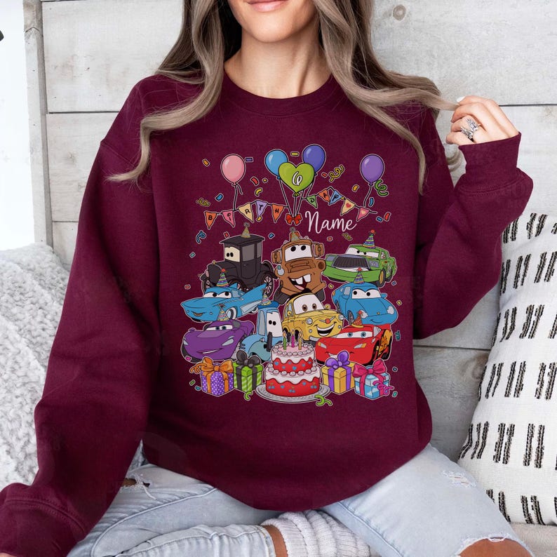 Peut inclure: Sweat-shirt bordeaux avec un motif color&eacute; de voitures de dessins anim&eacute;s, un g&acirc;teau d'anniversaire, des ballons et des cadeaux emball&eacute;s. Le mot "Name" est &eacute;crit en script au-dessus des voitures. Le design est festif et ludique.