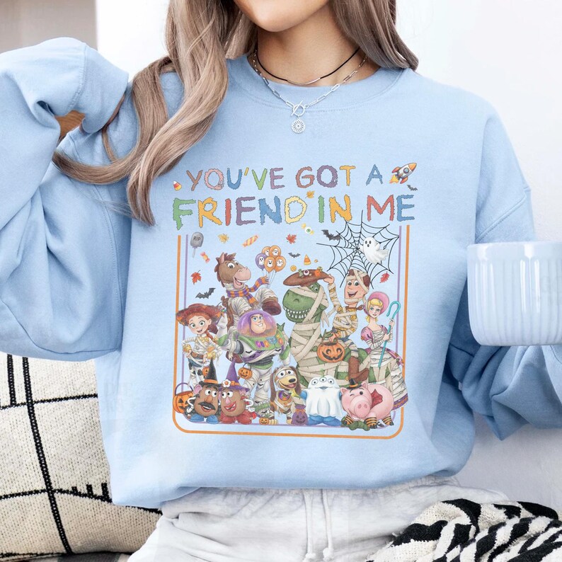 K&ouml;nnte beinhalten: Ein hellblaues Sweatshirt mit Rundhalsausschnitt und dem Text "YOU'VE GOT A FRIEND IN ME" &uuml;ber einer grafischen Illustration verschiedener Zeichentrickfiguren in Halloween-Kost&uuml;men, darunter eine Mumie, ein Geist und ein K&uuml;rbiseimer.