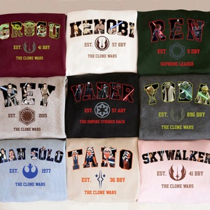 May include: Nine sweatshirts with Star Wars character names in a collegiate style. The sweatshirts are in various colors, including maroon, white, green, gray, black, light blue, beige, and pink. Each sweatshirt features a character name, the phrase "EST." and "The Clone Wars".