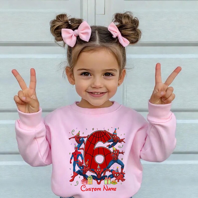 May include: A light pink long-sleeve sweatshirt featuring a large red number six graphic surrounded by multiple Spider-Man characters wearing party hats, colourful confetti, and gift boxes, with the text Custom Name printed at the bottom.