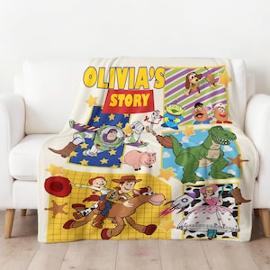 Op de afbeelding: Een cr&egrave;mekleurige deken met personages uit de animatiefilm Toy Story. De deken toont Buzz Lightyear, Woody, Jessie en andere personages. De tekst "OLIVIA'S STORY" is gedrukt in geel en rood. De deken is over een witte bank gedrapeerd.