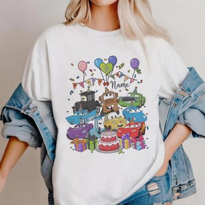 Peut inclure: T-shirt blanc avec un motif de dessin anim&eacute; color&eacute; de personnages de voitures, de ballons, de confettis et d'un g&acirc;teau d'anniversaire. Le motif comprend le mot "Name" sur une banni&egrave;re, sugg&eacute;rant un th&egrave;me d'anniversaire personnalis&eacute;.