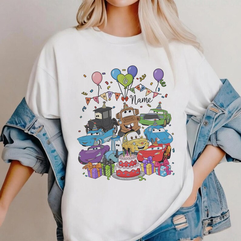 Peut inclure: T-shirt blanc avec un motif de dessin anim&eacute; color&eacute; de personnages de voitures, de ballons, de confettis et d'un g&acirc;teau d'anniversaire. Le motif comprend le mot "Name" sur une banni&egrave;re, sugg&eacute;rant un th&egrave;me d'anniversaire personnalis&eacute;.