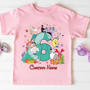 K&ouml;nnte beinhalten: Ein hellrosa T-Shirt mit kurzen &Auml;rmeln, das eine gro&szlig;e t&uuml;rkisfarbene Zahl sechs zeigt, umgeben von Zeichentrickfiguren wie einer Meerjungfrau, einem Prinzen, einer Meerhexe, einem Krebs und einem Hund mit Partyh&uuml;ten sowie dem Text Custom Name.