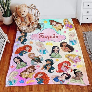 May include: A personalized blanket featuring various cartoon princesses in a colorful design. The blanket has a pink cloud with the name "Sophia" written in cursive. The princesses are surrounded by stars and crowns.
