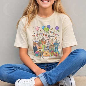 Puede incluir: Camiseta beige con un gr&aacute;fico de dibujos animados de varios personajes de animales, globos, una tarta de cumplea&ntilde;os y la palabra "Name". El dise&ntilde;o incluye confeti y una pancarta. La camiseta la lleva una persona con vaqueros azules.