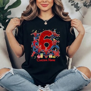 May include: A black short-sleeve t-shirt featuring a large red number six with a spiderweb pattern. The design includes multiple illustrations of Spider-Man wearing party hats, colourful gift boxes, and the text Custom Name in red font.