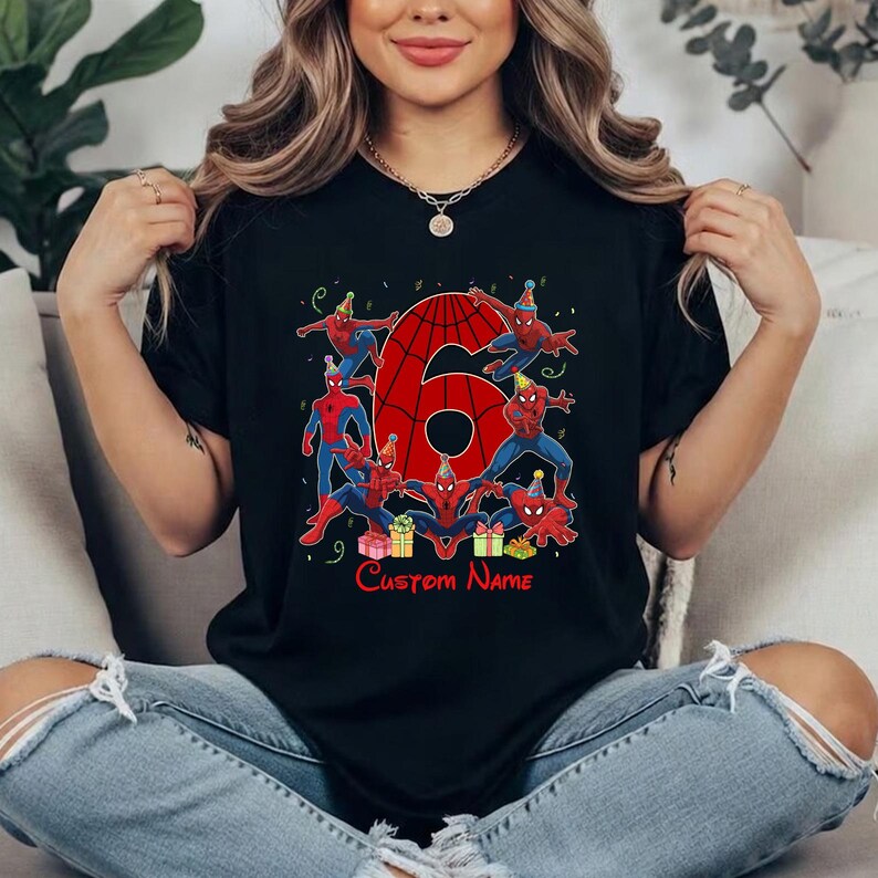 May include: A black short-sleeve t-shirt featuring a large red number six with a spiderweb pattern. The design includes multiple illustrations of Spider-Man wearing party hats, colourful gift boxes, and the text Custom Name in red font.