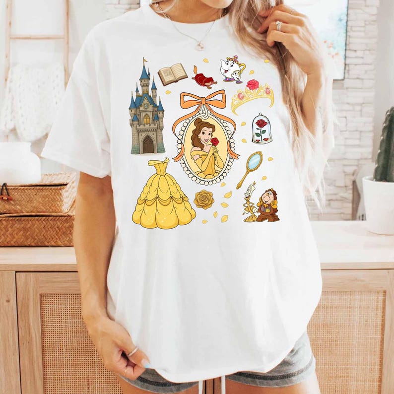 May include: White t-shirt featuring a collection of cartoon illustrations. The design includes a yellow ball gown, a castle, a portrait of a woman, a rose, and other themed elements. The shirt is a casual, everyday wear item.