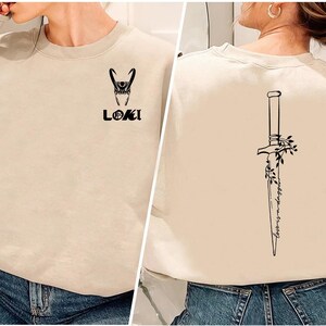 May include: Beige sweatshirt featuring a black graphic of Loki's helmet and the word "LOKI" on the front. The back displays a black dagger design with floral accents and the text "lovers & dagger".
