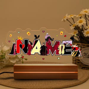 May include: A personalized night light featuring the word "NAME" with Disney villains. The letters are in black, red, and yellow, with character illustrations. The light sits on a wooden base and is plugged in.