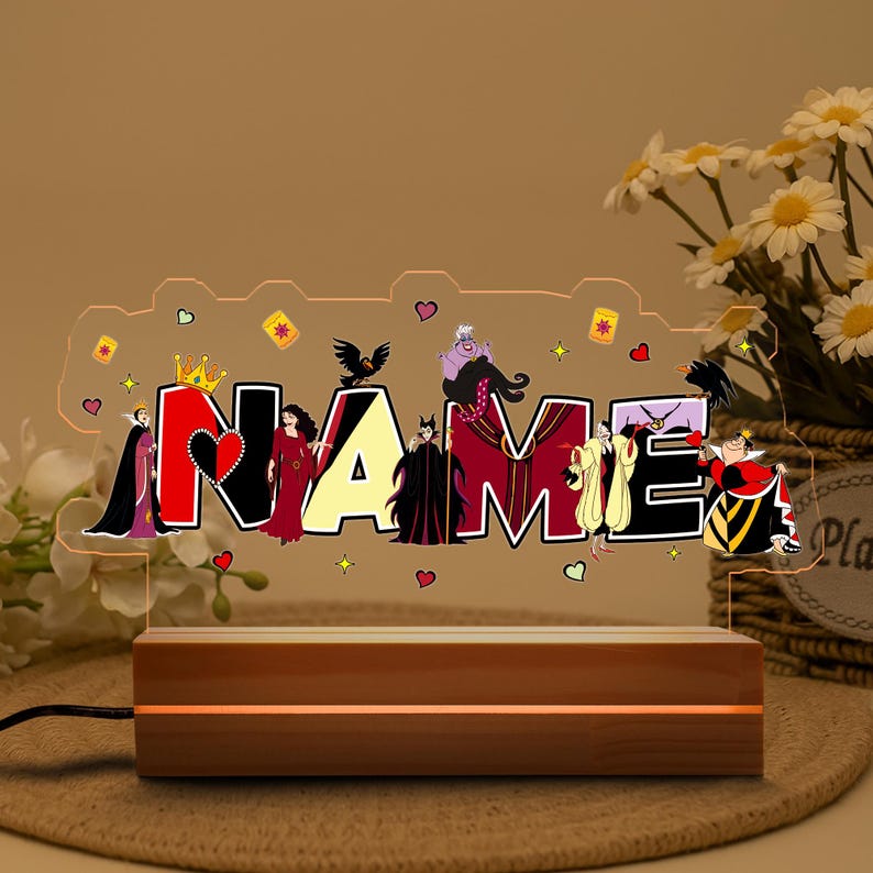 May include: A personalized night light featuring the word "NAME" with Disney villains. The letters are in black, red, and yellow, with character illustrations. The light sits on a wooden base and is plugged in.
