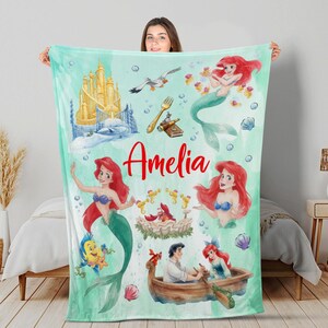 May include: A teal blanket with illustrations of a mermaid, a castle, and other characters. The name "Amelia" is written in red script. The blanket has a watercolour style with bubbles and seashell accents.
