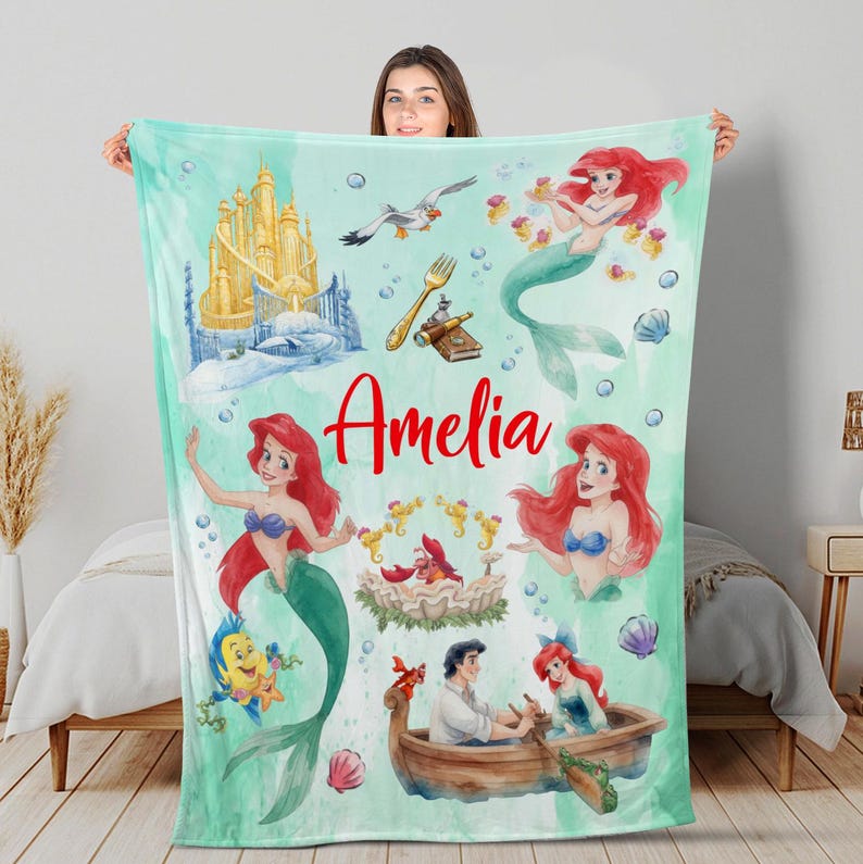May include: A teal blanket with illustrations of a mermaid, a castle, and other characters. The name "Amelia" is written in red script. The blanket has a watercolour style with bubbles and seashell accents.
