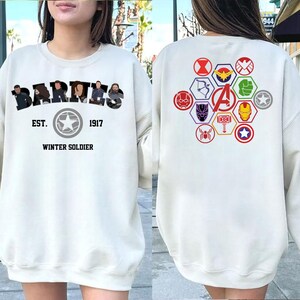 Puede incluir: Sudadera color crema con la palabra "BARTES" sobre "EST. 1917 WINTER SOLDIER" y un emblema de estrella. La parte trasera muestra s&iacute;mbolos de superh&eacute;roes y la letra "A" en un hex&aacute;gono, en un dise&ntilde;o colorido.