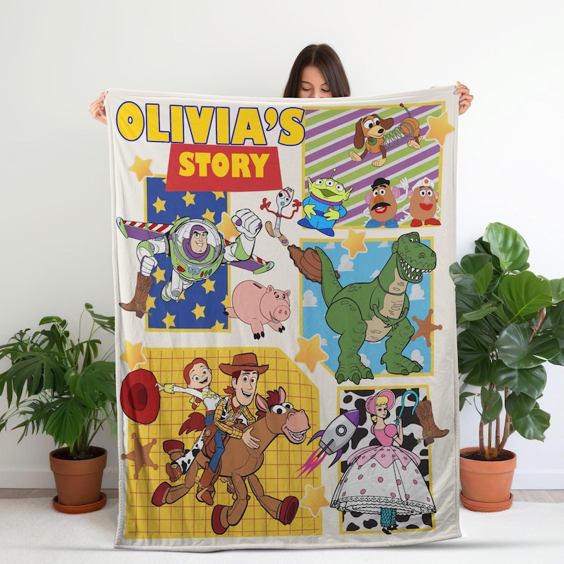Op de afbeelding: Een cr&egrave;mekleurige deken met een patchwork-ontwerp met Toy Story-personages. De deken bevat Buzz Lightyear, Woody, Jessie, Rex en anderen, met de tekst "OLIVIA'S STORY" in geel en rood.
