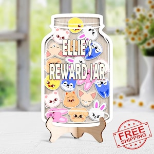May include: A wooden reward jar with the text "ELLIE'S REWARD JAR" and various cartoon animal stickers. The jar is displayed on a wooden stand. The background is a blurred view of a window and flowers.