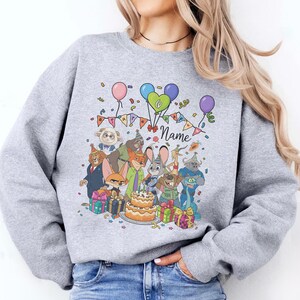 Puede incluir: Una sudadera gris claro con una colorida ilustraci&oacute;n de dibujos animados de personajes, globos, confeti y una tarta de cumplea&ntilde;os. El dise&ntilde;o incluye la palabra "Name" y el n&uacute;mero "6".