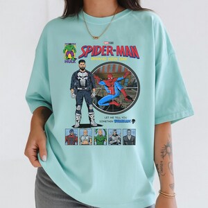 May include: A light blue t-shirt featuring a comic book style graphic of Spider-Man, The Punisher, and other Marvel characters. The shirt has the text "SPIDER-MAN BRAND NEW DAY" and "LET ME TELL YOU SOMETHIN WEBBHEAD!"