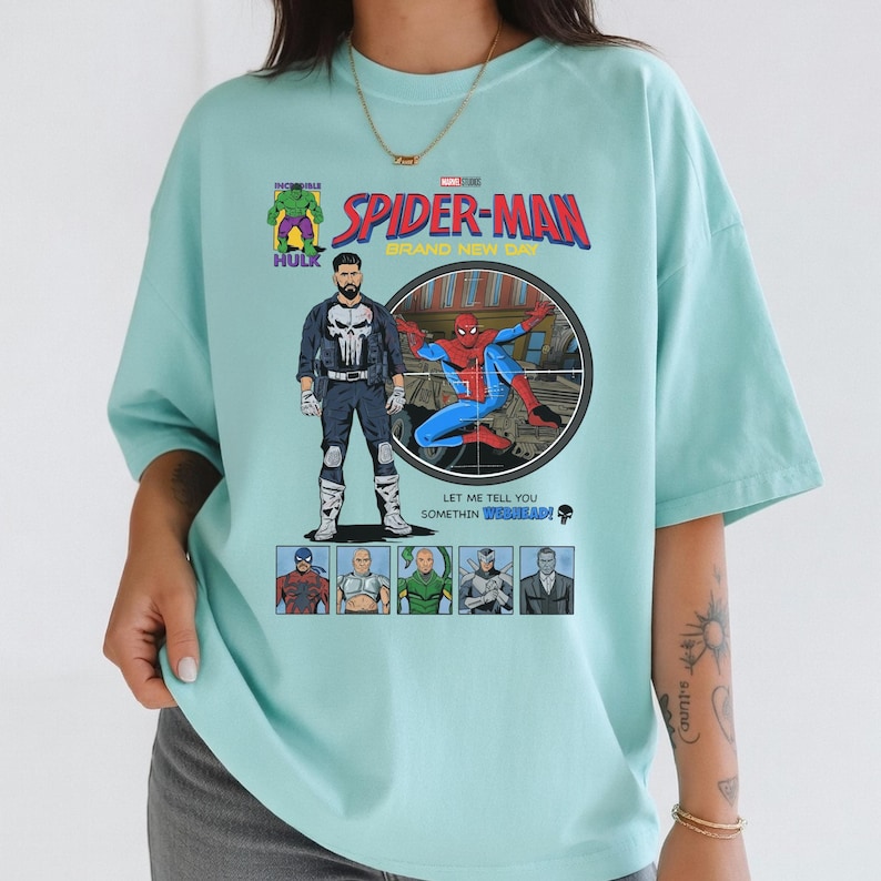 May include: A light blue t-shirt featuring a comic book style graphic of Spider-Man, The Punisher, and other Marvel characters. The shirt has the text "SPIDER-MAN BRAND NEW DAY" and "LET ME TELL YOU SOMETHIN WEBBHEAD!"