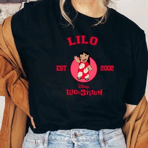 May include: Black t-shirt featuring a graphic of Lilo from Disney's Lilo & Stitch. The design includes the word "LILO" in red, the text "EST 2002", and the Disney logo. Lilo is depicted in a red dress with white spots, inside a pink circle.