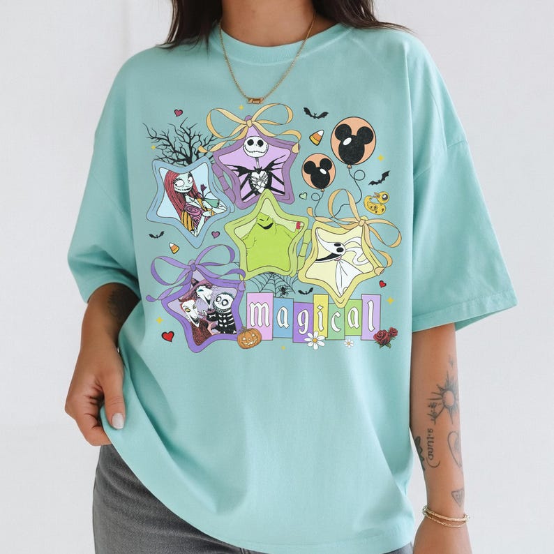 May include: A light blue t-shirt with a colourful graphic design. The design features star-shaped gift boxes with characters, bats, and the word "magical". The shirt has short sleeves and a relaxed fit.