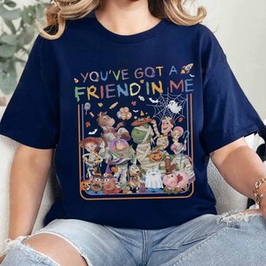 K&ouml;nnte beinhalten: Ein dunkelblaues T-Shirt mit einer bunten Grafik verschiedener Zeichentrickfiguren in Halloween-Kost&uuml;men. Der Text "YOU'VE GOT A FRIEND IN ME" ist &uuml;ber den Figuren aufgedruckt, begleitet von kleinen Symbolen wie S&uuml;&szlig;igkeiten, Flederm&auml;usen und einem Spinnennetz.