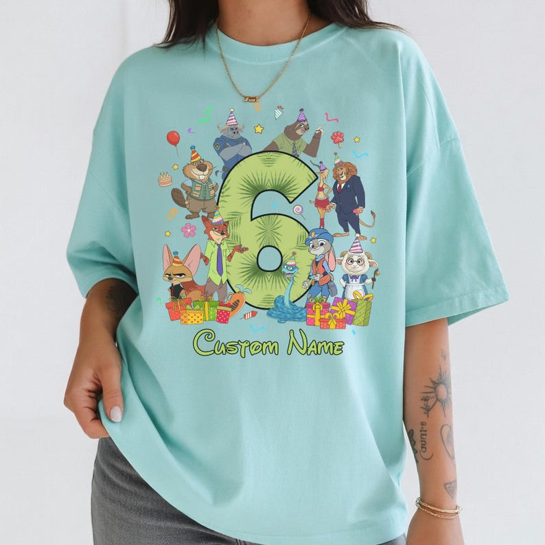 May include: A light blue t-shirt featuring a large green number six surrounded by various animated animal characters wearing party hats. The graphic includes colorful gift boxes, confetti, and the text Custom Name printed in green below the design.