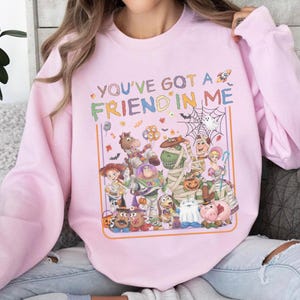 K&ouml;nnte beinhalten: Ein hellrosa Sweatshirt mit Rundhalsausschnitt und einer bunten Grafik verschiedener Zeichentrickfiguren in Halloween-Kost&uuml;men. Der Text "YOU'VE GOT A FRIEND IN ME" ist &uuml;ber den Figuren aufgedruckt, begleitet von kleinen Symbolen wie S&uuml;&szlig;igkeiten, Flederm&auml;usen und einem Spinnennetz.
