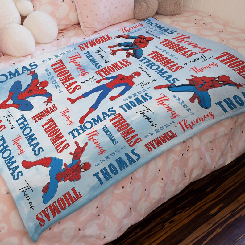 May include: A light blue blanket featuring multiple illustrations of Spider-Man in various action poses. The blanket has the name "Thomas" printed repeatedly in red and blue, creating a personalized design.